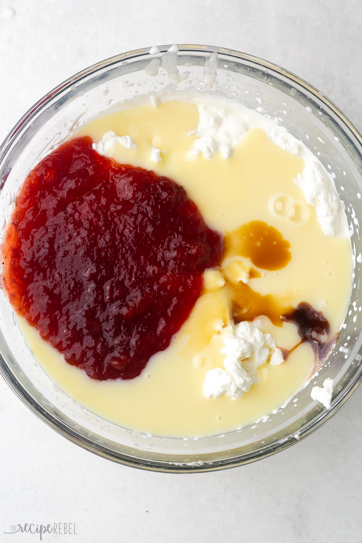 Top view of glass mixing bowl with whipped cream in it topped with strawberry mix, vanilla, and sweetened condensed milk.