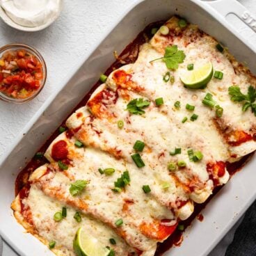 overhead view of a white baking dish filled with beef enchiladas and toppings beside.