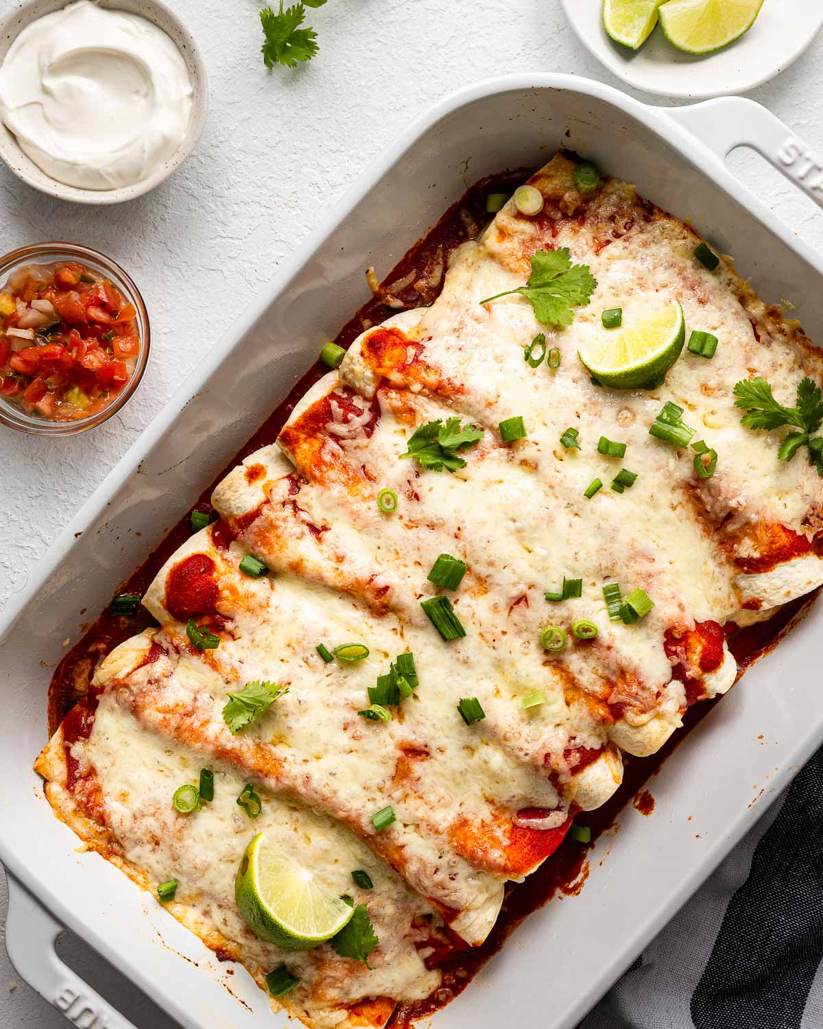 overhead view of a white baking dish filled with beef enchiladas and toppings beside.