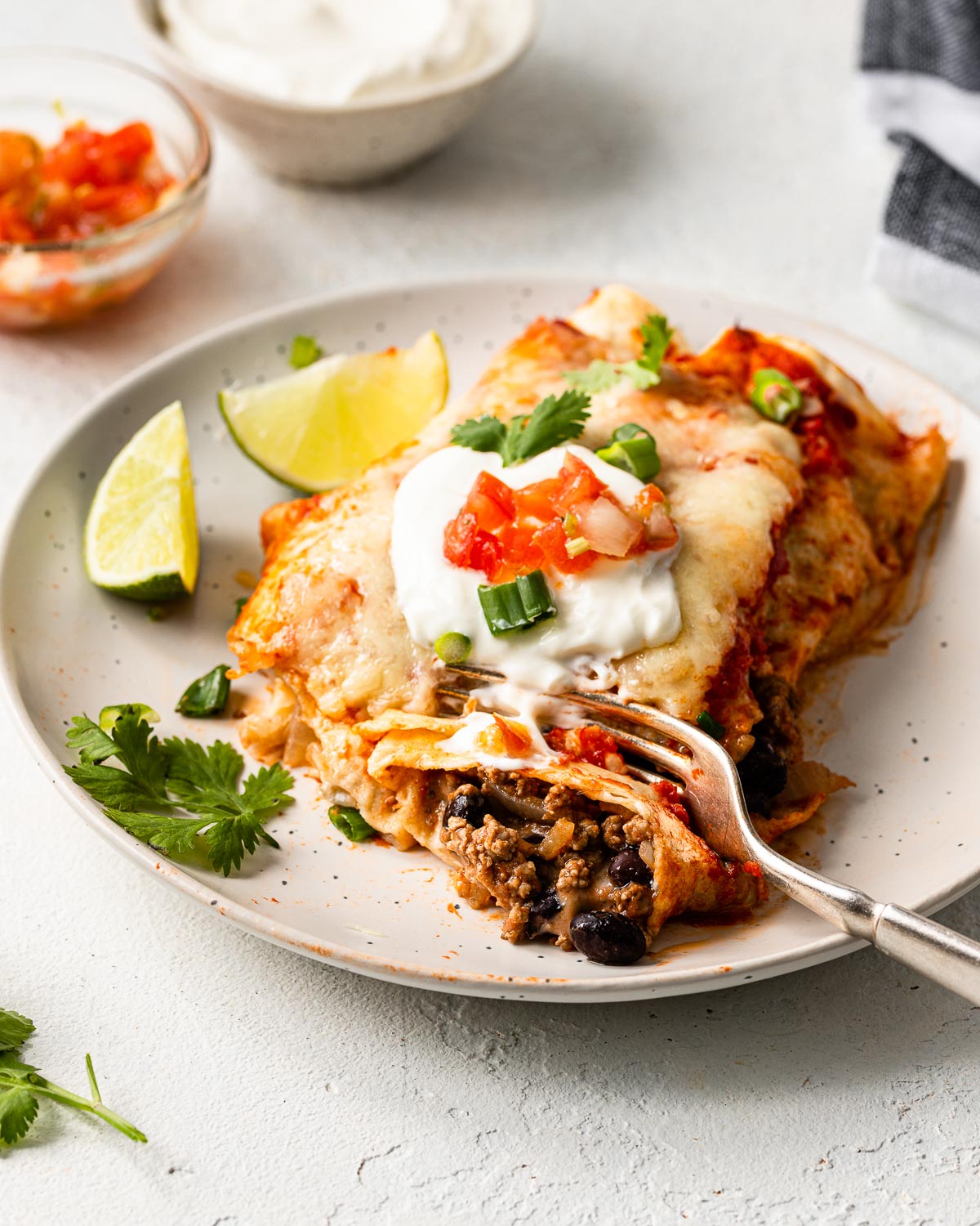 close up of a plate beef enchiladas with a fork in it and toppings in the background.