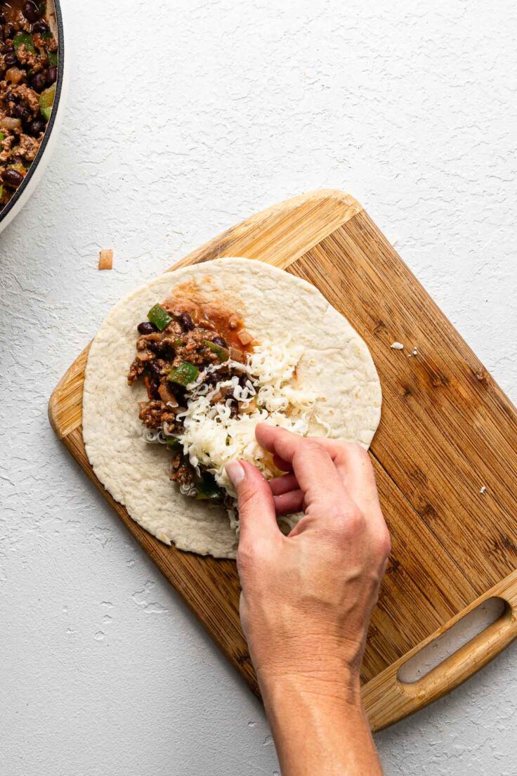 a hand adding shredded cheese to a beef taquito on a cutting board.