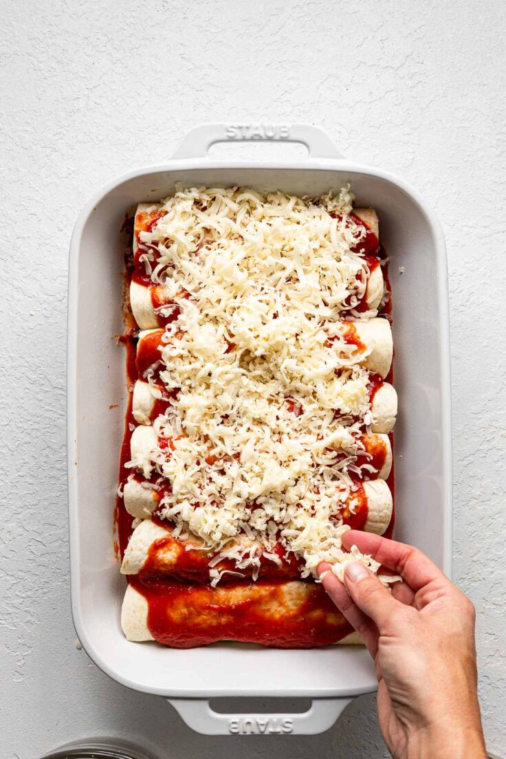 white pan filled with beef taquitos being topped with shredded cheese.
