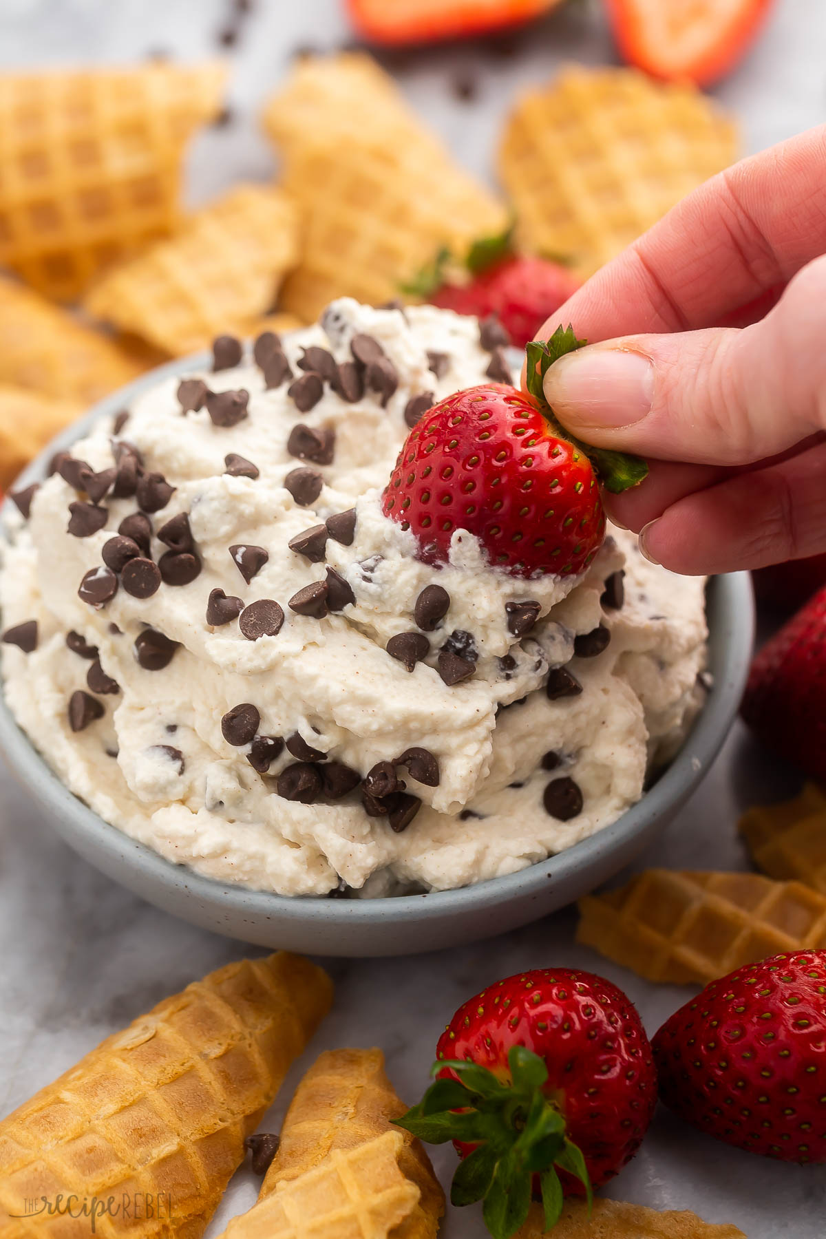 a strawberry being dipped into a bowl of cannoli dip with waffle cones and strawberries beside.