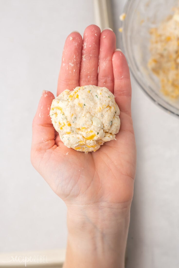 a hand holding an unbaked rolled biscuit by a mixing bowl.