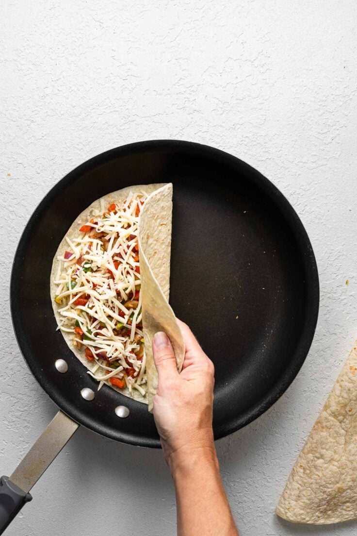 a hand folding together a cheese quesadilla in a black frying pan.