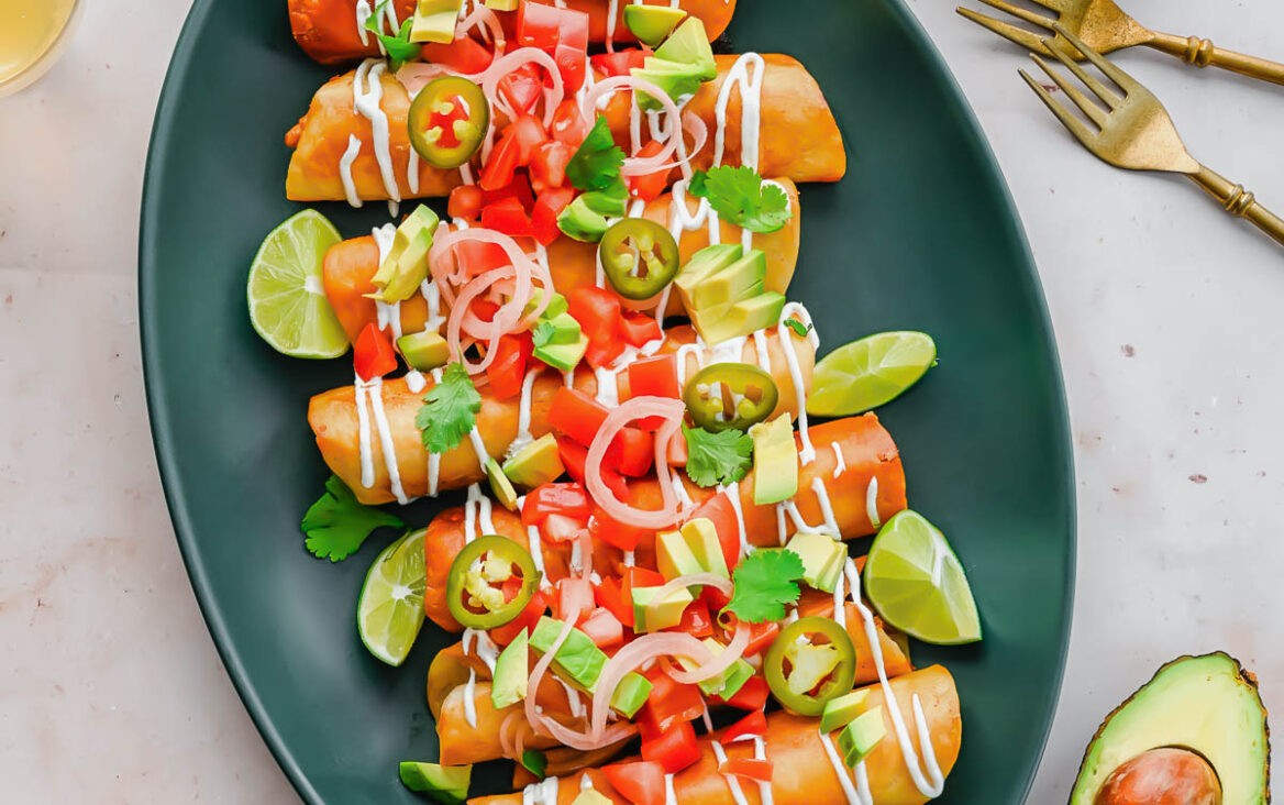 close up of a platter of chicken taquitos with toppings on a grey surface.