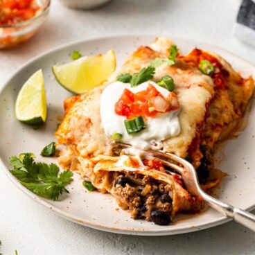 close up of a plate beef enchiladas with a fork in it and toppings in the background.