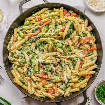 big black pan filled with cooked pasta primavera with glass of water and vegetables beside.