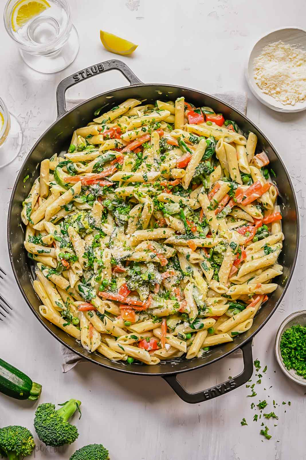 big black pan filled with cooked pasta primavera with glass of water and vegetables beside.