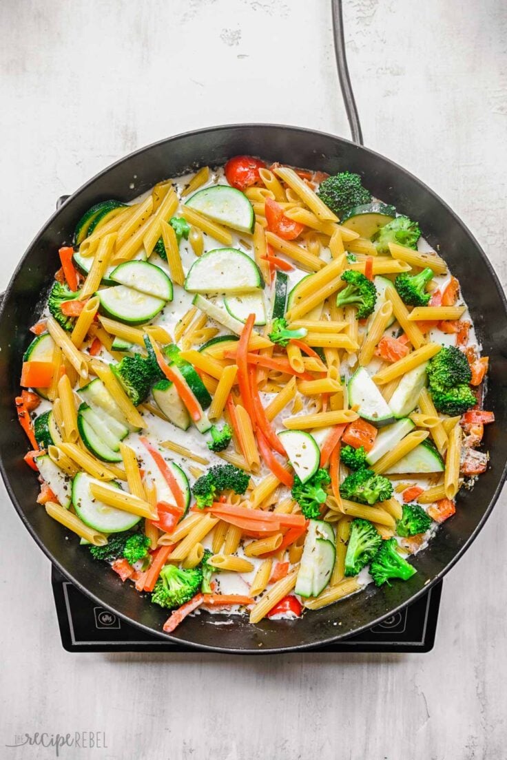 black frying pan with pasta and vegetables added on top of cream sauce.