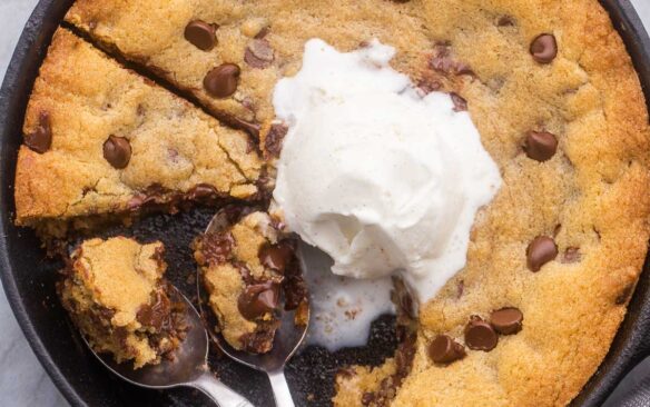 overhead close up of skillet cookie in pan with spoons and vanilla ice cream on top.