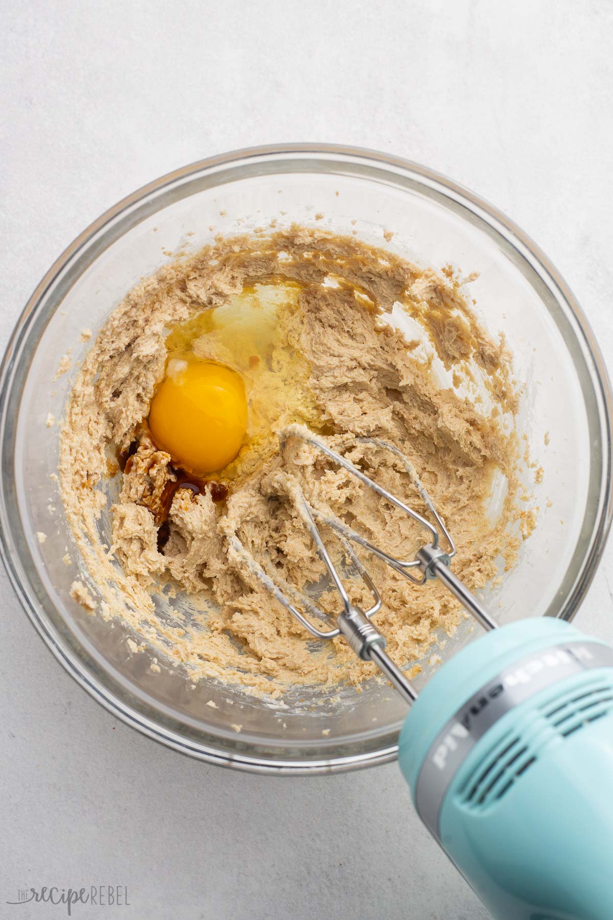 mixed ingredients in glass bowl with egg added and mixer beside.