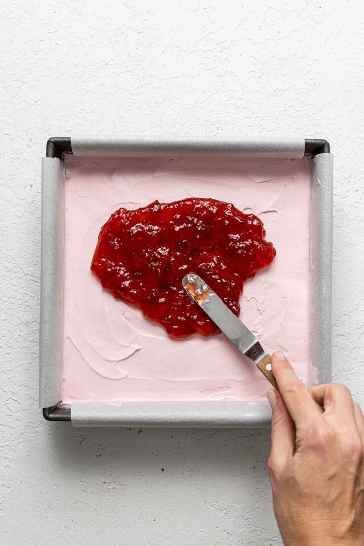 spreading strawberry jam on top of the strawberry ice cream in cake pan.