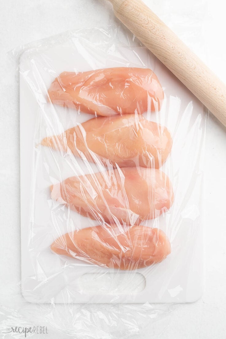 four chicken breasts lying on a cutting board covered in plastic wrap with a wooden rolling pin beside.