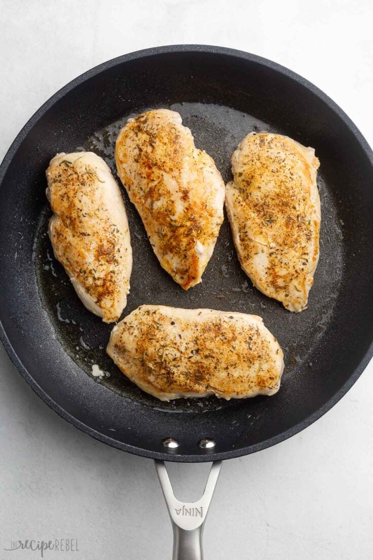 four cooked and seasoned chicken breasts lying in a black frying pan.