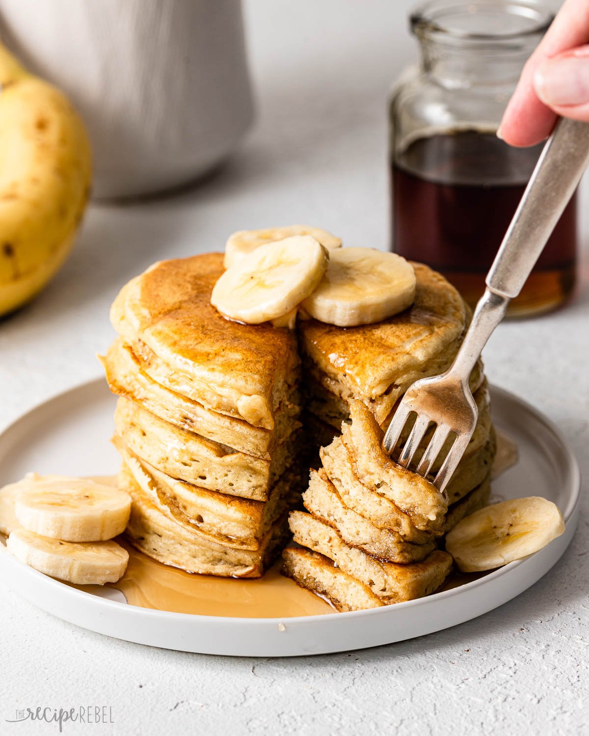 a fork full of pancakes with a stack of pancakes, bananas, and syrup.