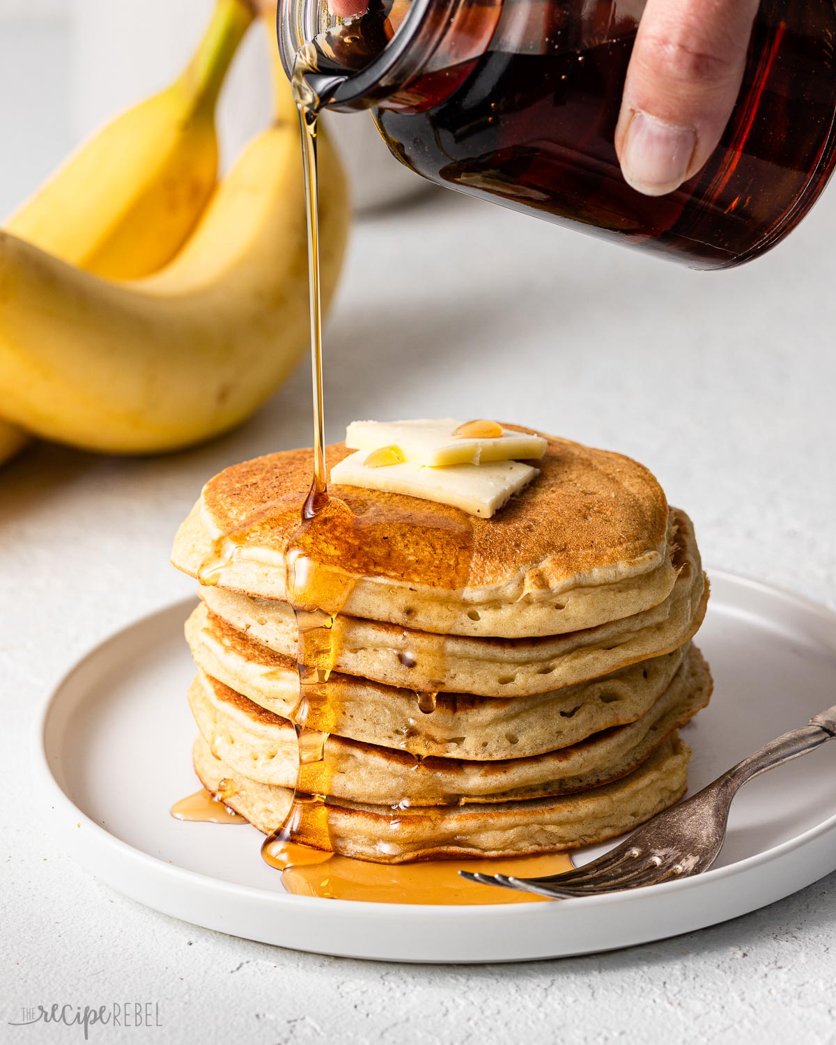 a stack of pancakes on a white plate with syrup being poured on top.