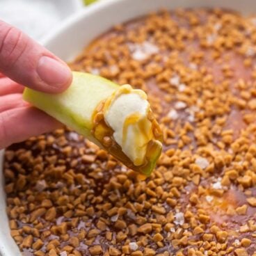 a hand holding an apple slice with caramel apple dip on it.