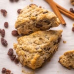 two scones on parchment paper with raisins and cinnamon sticks beside.