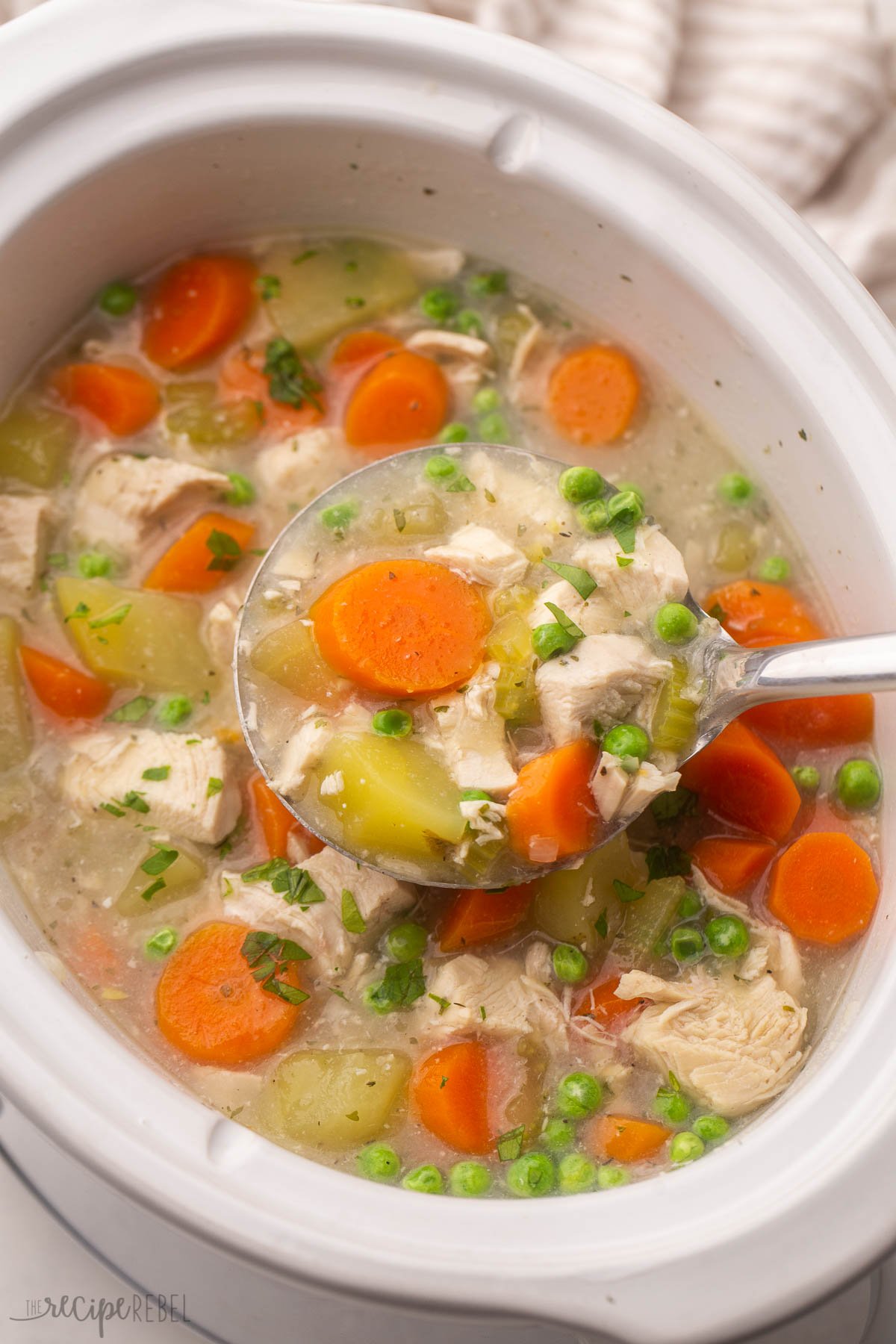 overhead shot of a white crockpot full of chicken stew with a steel ladle scooping out.