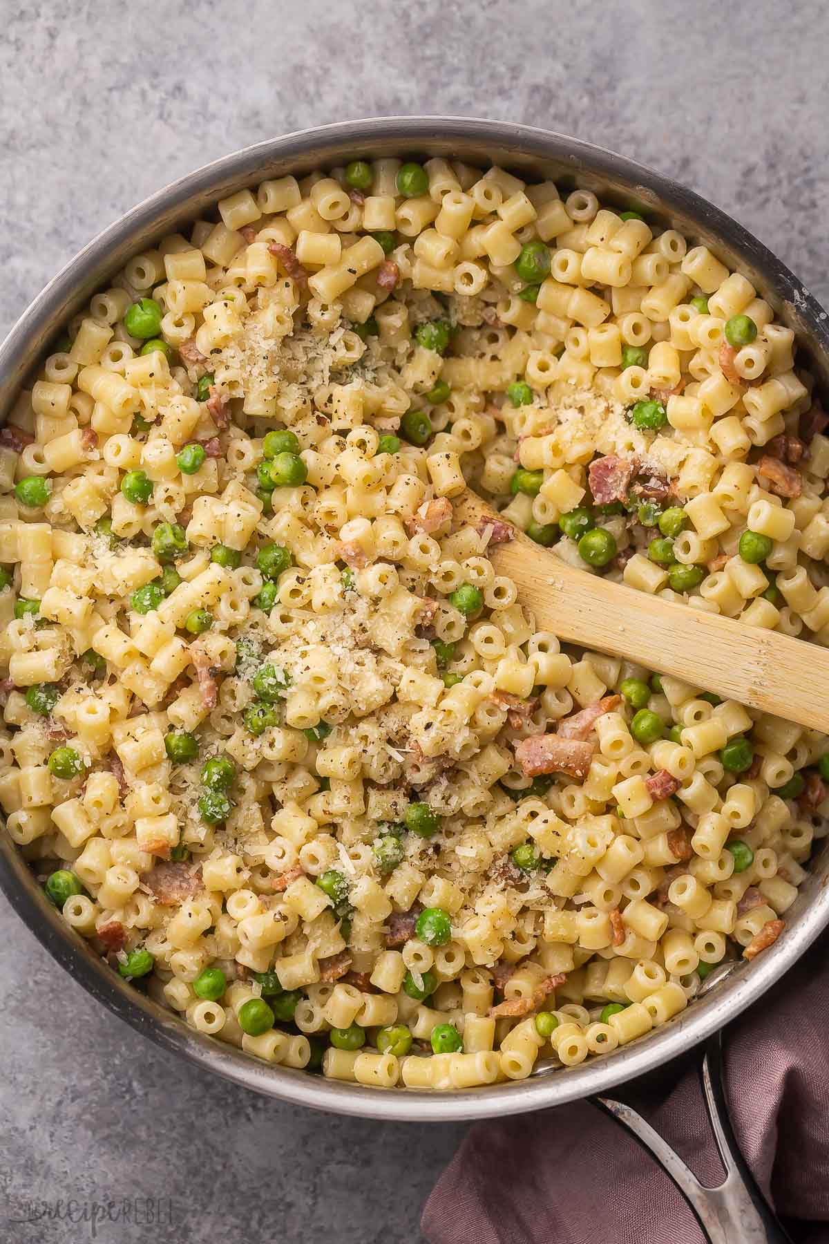 large steel pan filled with ditalini pasta, bacon, peas and a wooden ladle.