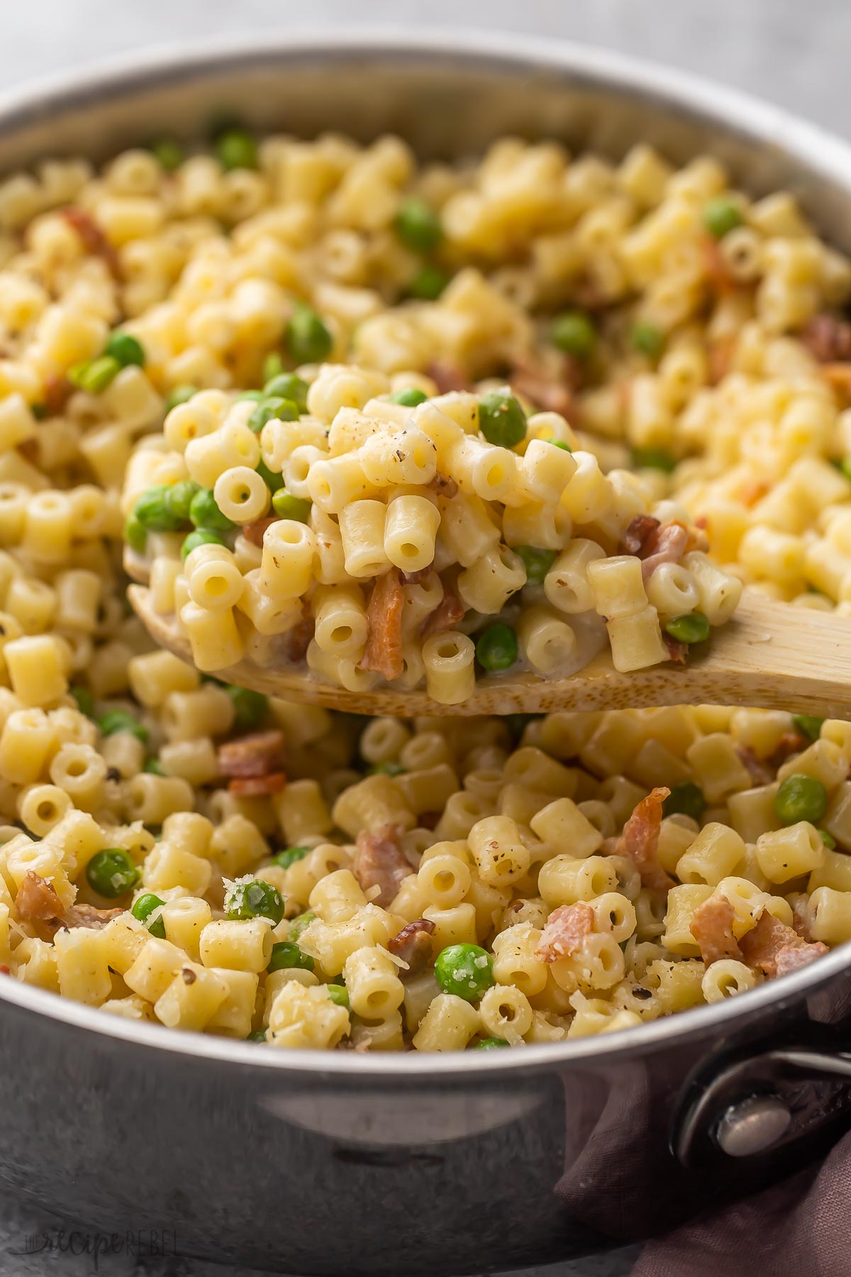 ditalini pasta with bacon and peas being scooped out of pan in wooden ladle.