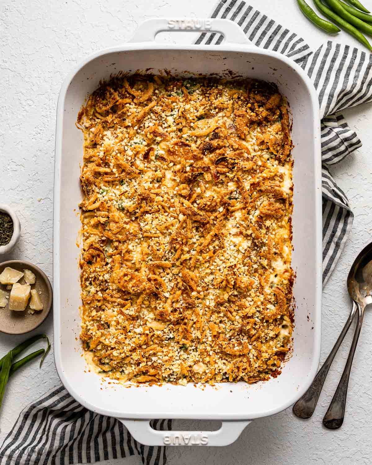 white baking dish full of baked green bean casserole surrounded by spoons, cheese and green beans.