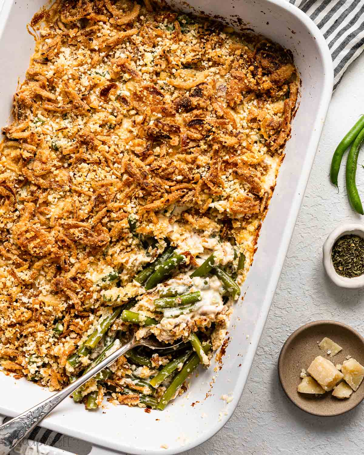 a spoon lying in a white pan of green bean casserole on a white surface.