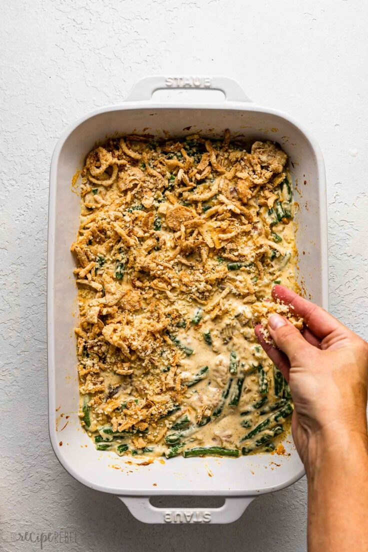 baked green bean casserole being topped with bread crumbs and fried onions.