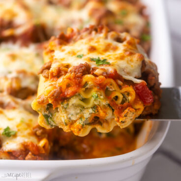 close up of a piece of lasagna roll up being scooped from a white baking dish.