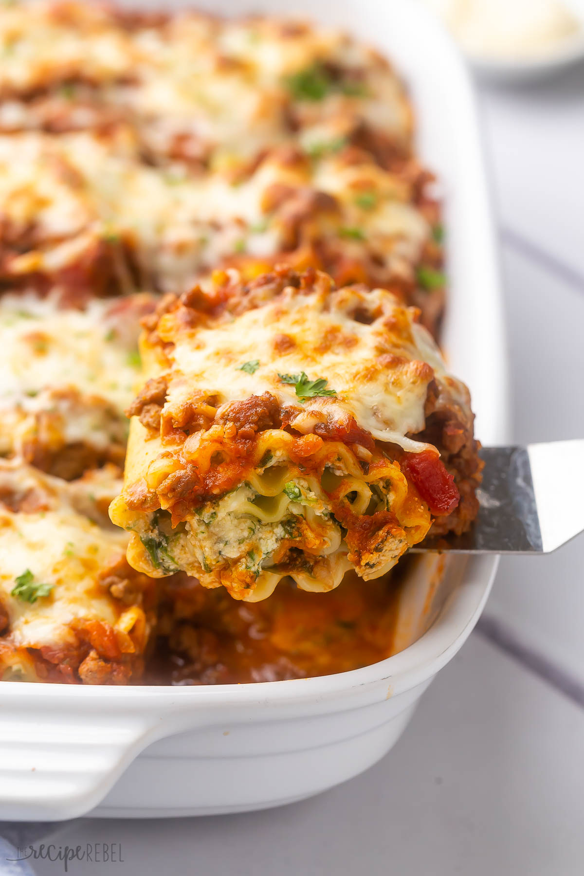 a steel spatula scooping a piece of lasagna roll up out of a white baking dish.