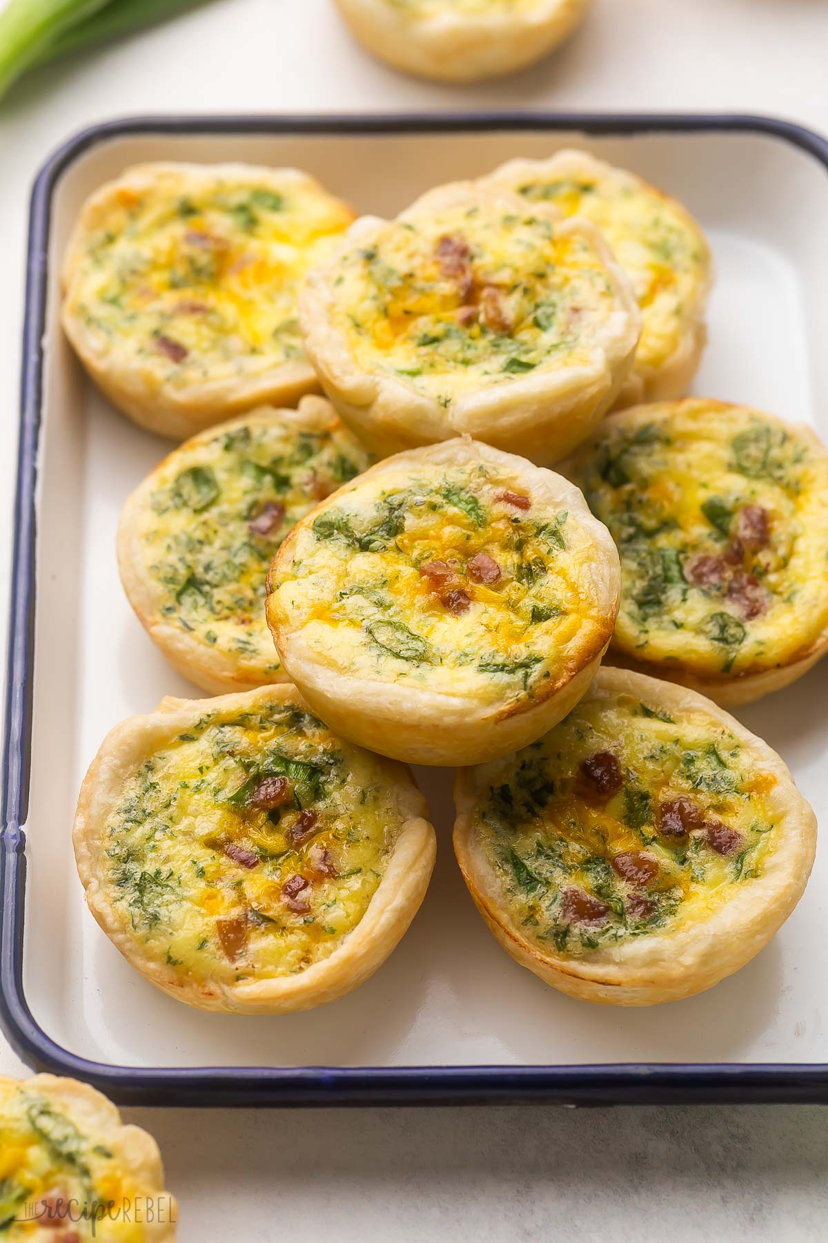 baked mini quiche stacked in white dish with mini quiche in background.