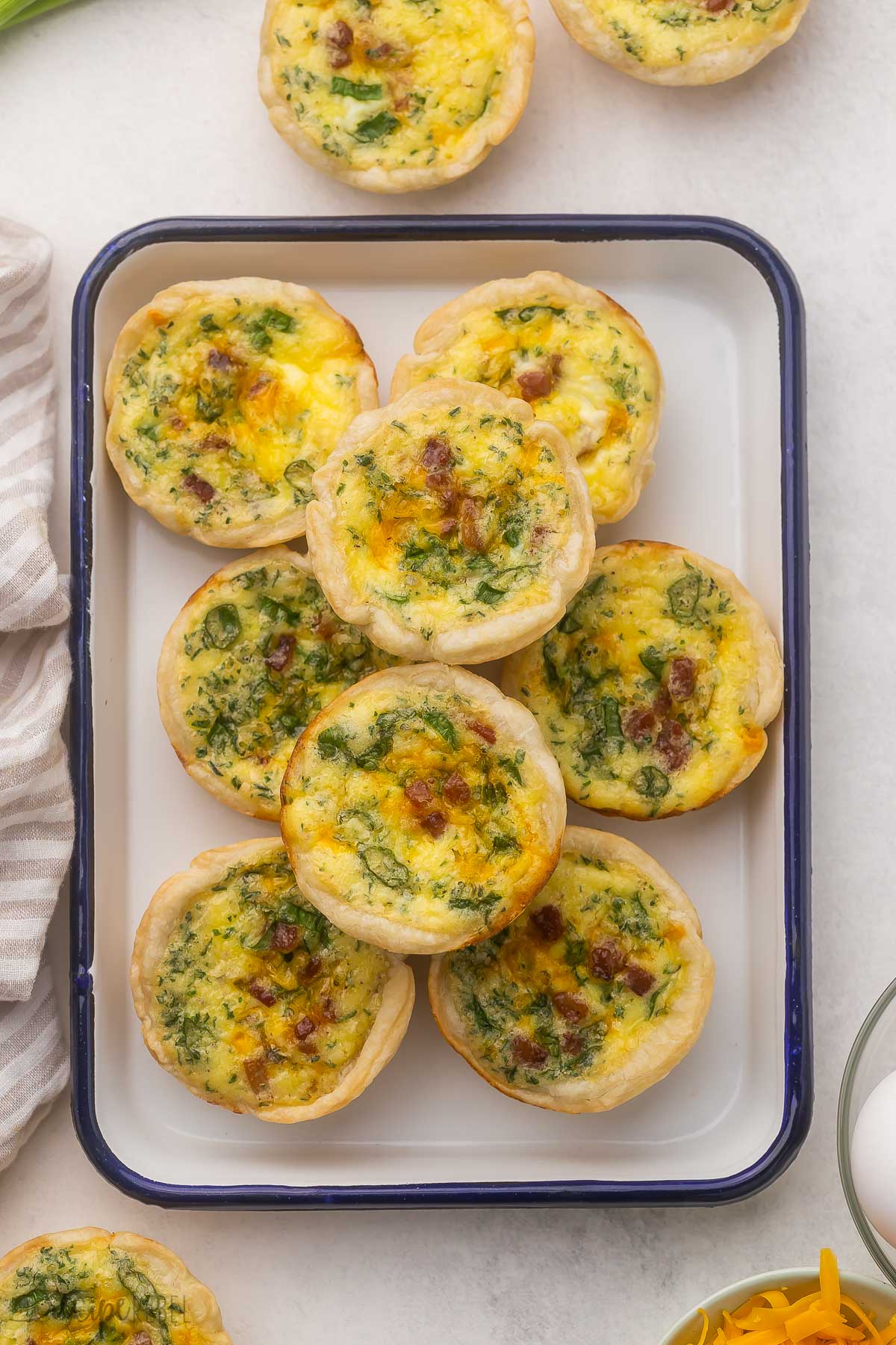 This Mini Quiche recipe is perfect for prepping ahead for a grab-and-go breakfast, to have as part of a breakfast or brunch spread, or as an appetizer at the holidays! They're super easy to make, and you can easily change their flavor with a few simple twists. #quiche #appetizer | holiday appetizers | brunch recipes | breakfast ideas | healthy recipes | bacon | spinach | pie crust | easy recipes overhead view of mini quiche in blue and white dish on grey surface.
