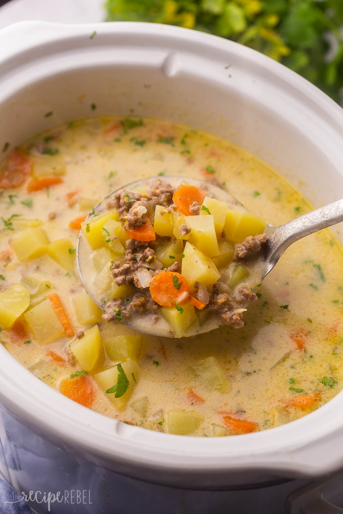 a full scoop of slow cooker cheeseburger soup above white slow cooker with parsley in the background.