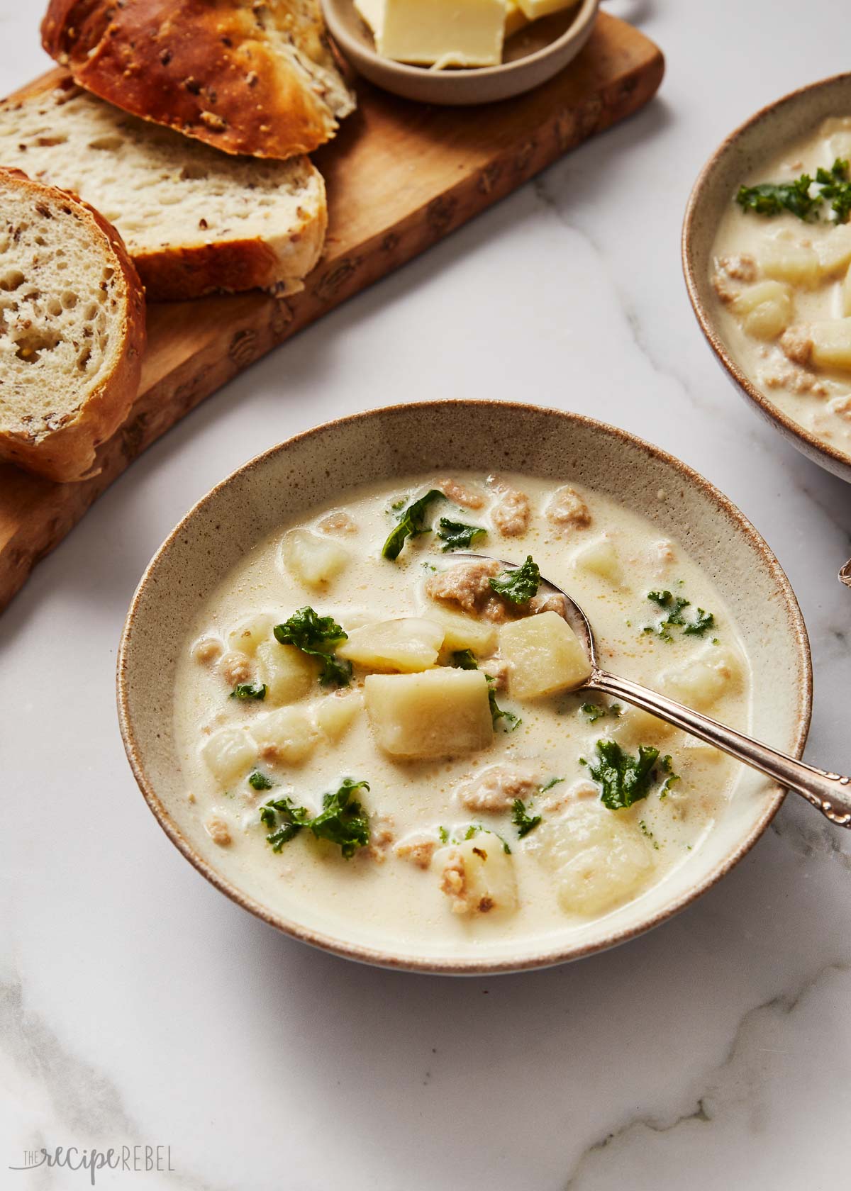 This easy Slow Cooker Zuppa Toscana is a creamy Tuscan soup full of Italian sausage, kale, potatoes, and Parmesan. It's hearty, creamy, and perfect for the colder months! There's a step-by-step recipe video down in the recipe card, too! #soup #slowcooker #crockpot | soup recipes | dinner ideas | easy dinner recipes | crockpot recipes | slow cooker meals a filled bowl of zuppa toscana soup with sliced bread beside.
