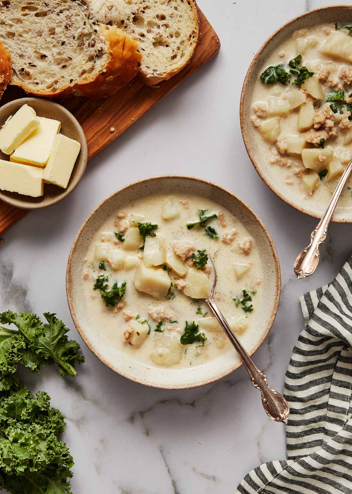 This easy Slow Cooker Zuppa Toscana is a creamy Tuscan soup full of Italian sausage, kale, potatoes, and Parmesan. It's hearty, creamy, and perfect for the colder months! There's a step-by-step recipe video down in the recipe card, too! #soup #slowcooker #crockpot | soup recipes | dinner ideas | easy dinner recipes | crockpot recipes | slow cooker meals overhead shot of bowls of slow cooker zuppa toscana with bread and butter beside.