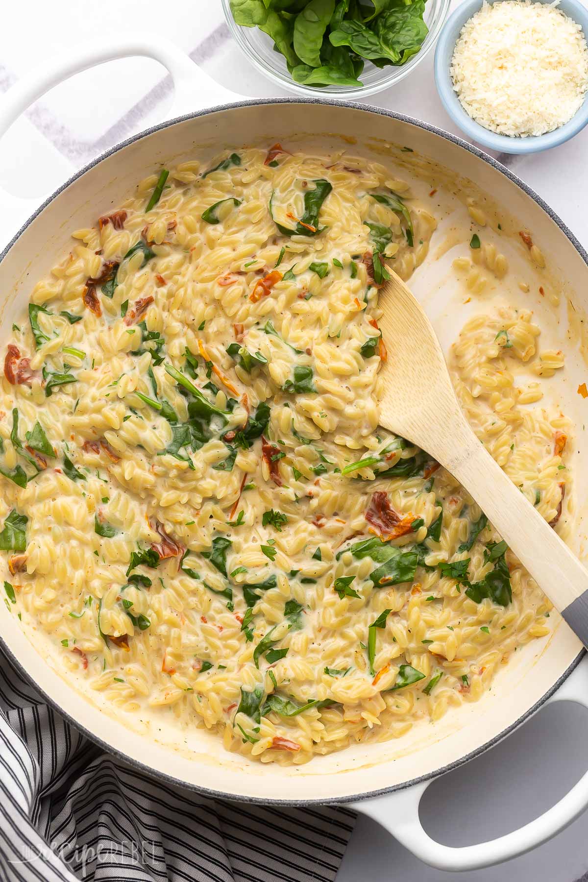 overhead of a white dish of sun dried tomato spinach orzo and wooden ladle with spinach and parmesan beside it.