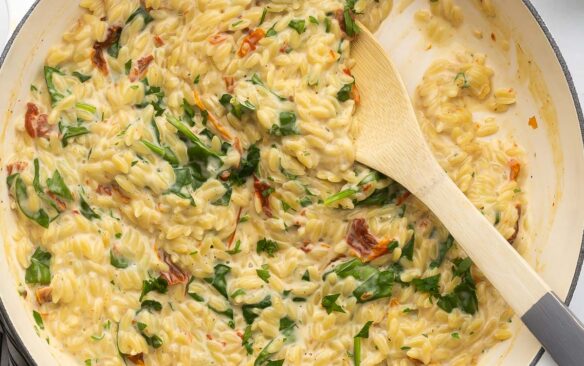 close up of a wooden ladle scooping sun dried tomato spinach orzo out of dish.