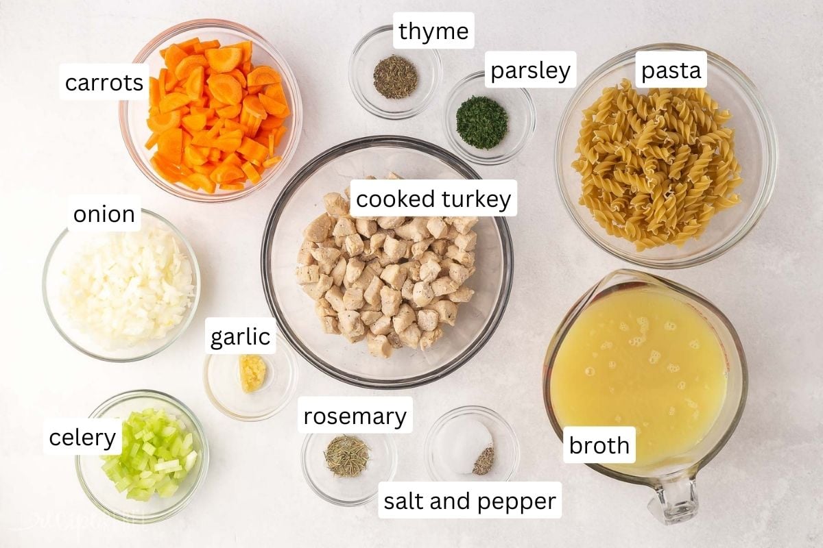 ingredients for turkey noodle soup in glass bowls on a grey surface.