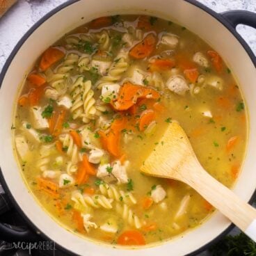 close up of wooden ladle in pot of turkey noodle soup with parsley sprinkled on top.