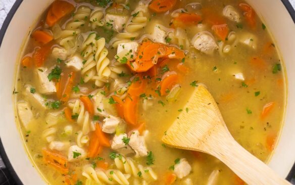 close up of wooden ladle in pot of turkey noodle soup with parsley sprinkled on top.