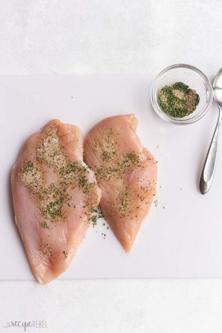 chicken breasts being seasoned on a cutting board.
