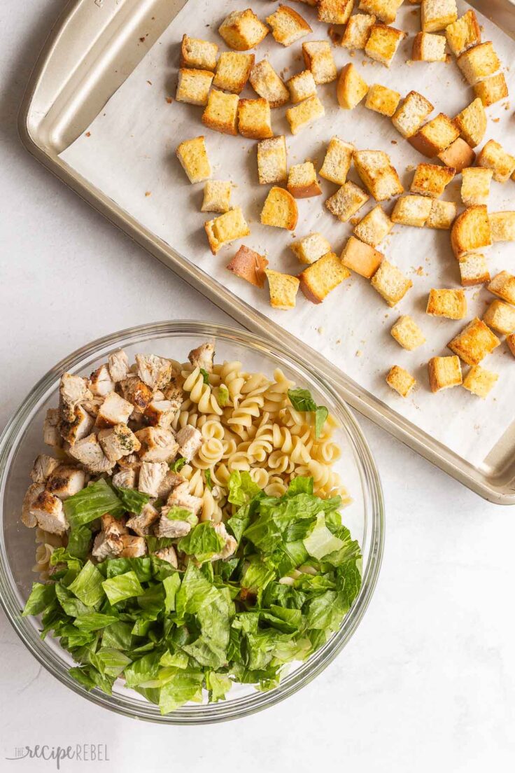 caesar pasta salad in a glass bowl with pan of croutons ready to add in.