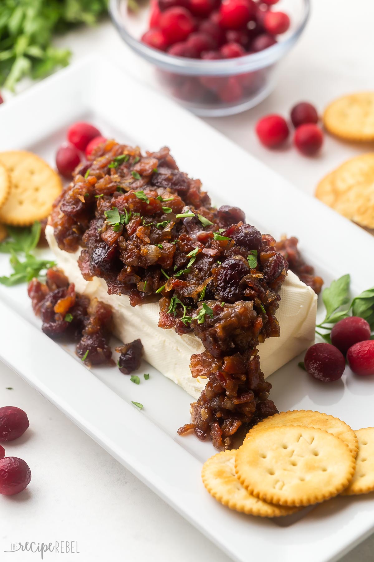 cream cheese on a white dish with cranberry bacon sauce, crackers, and cranberries in the background.