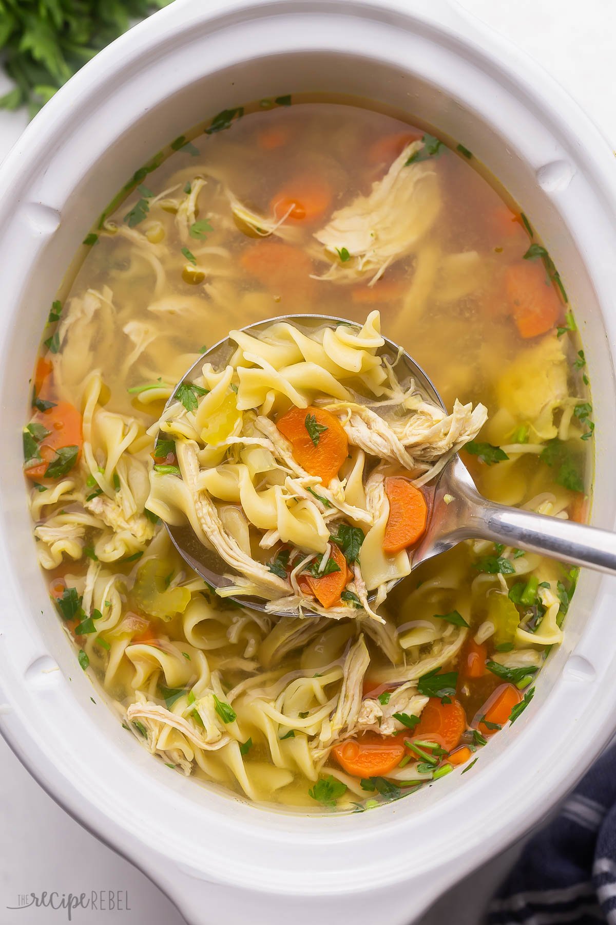 white crockpot full of chicken noodle soup with steel ladle scooping soup.