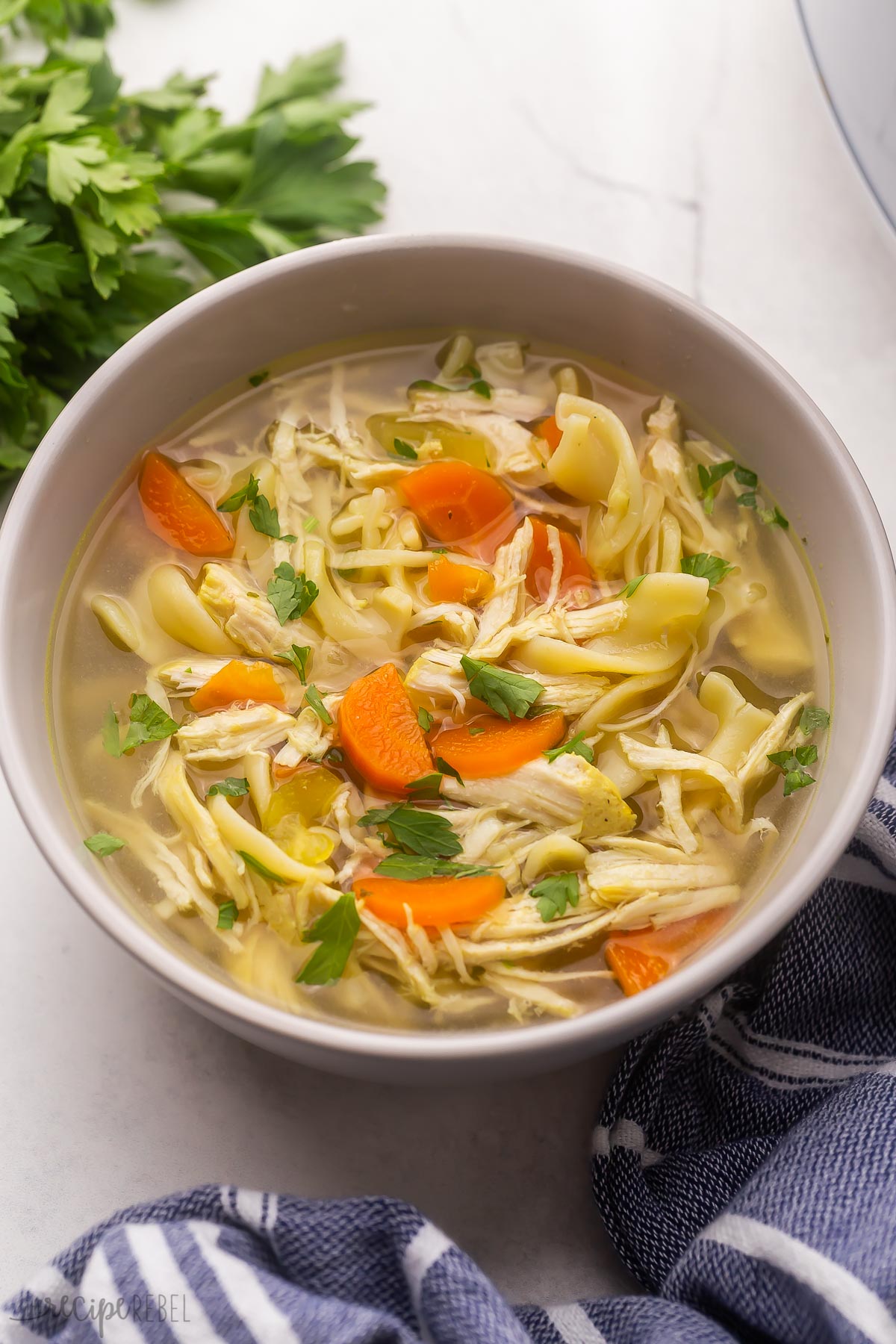 grey bowl of chicken noodle soup with fresh parsley in the background.