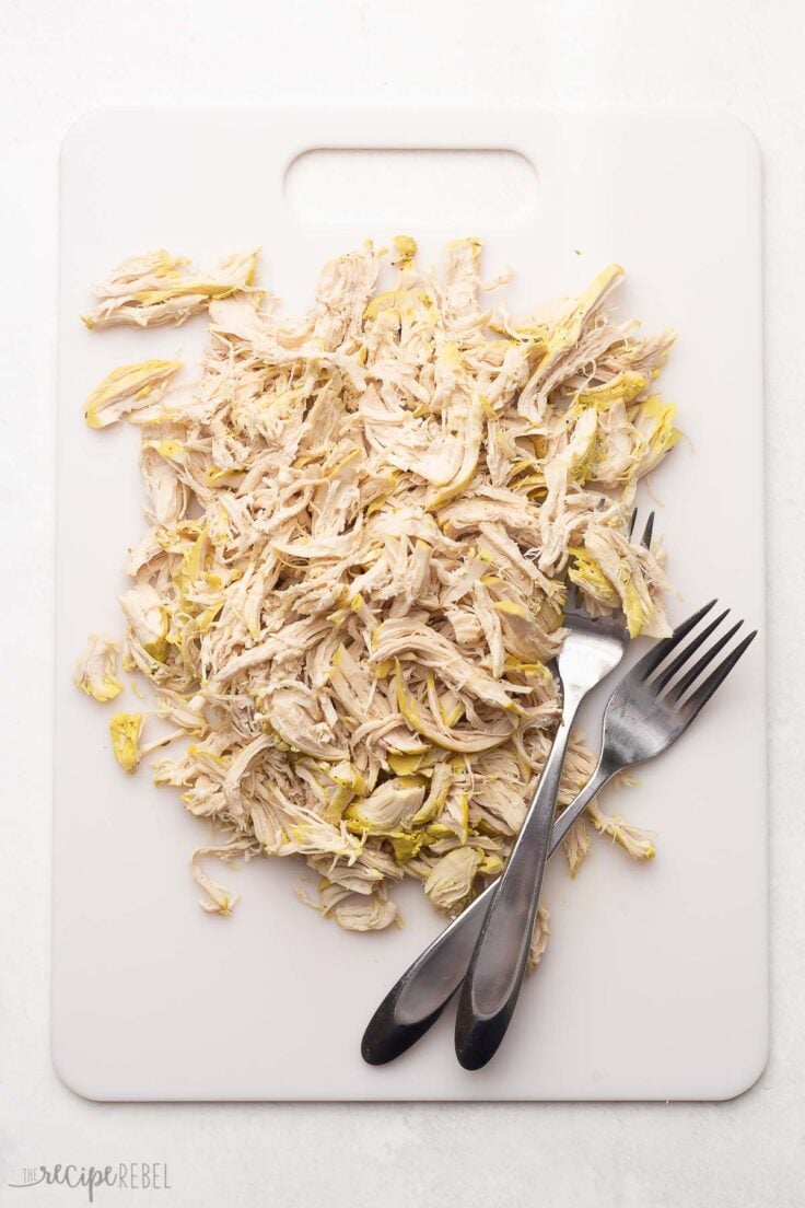 white cutting board with two forks and shredded chicken lying on it.