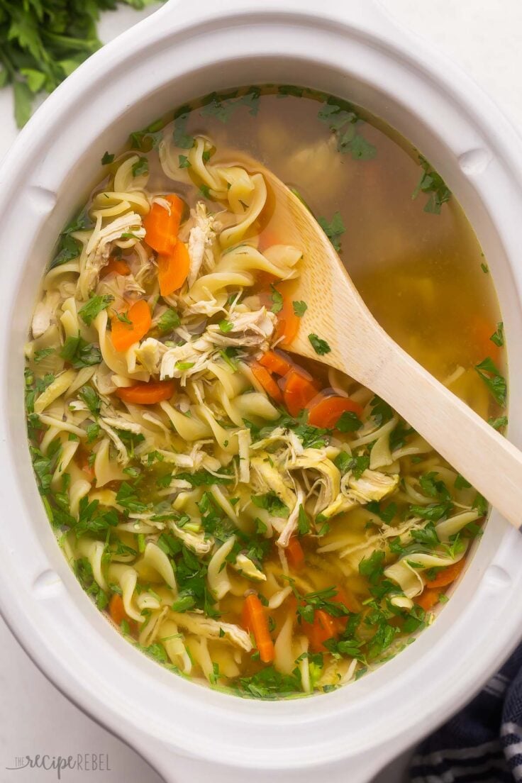 white crockpot full of chicken noodle soup with wooden ladle in.