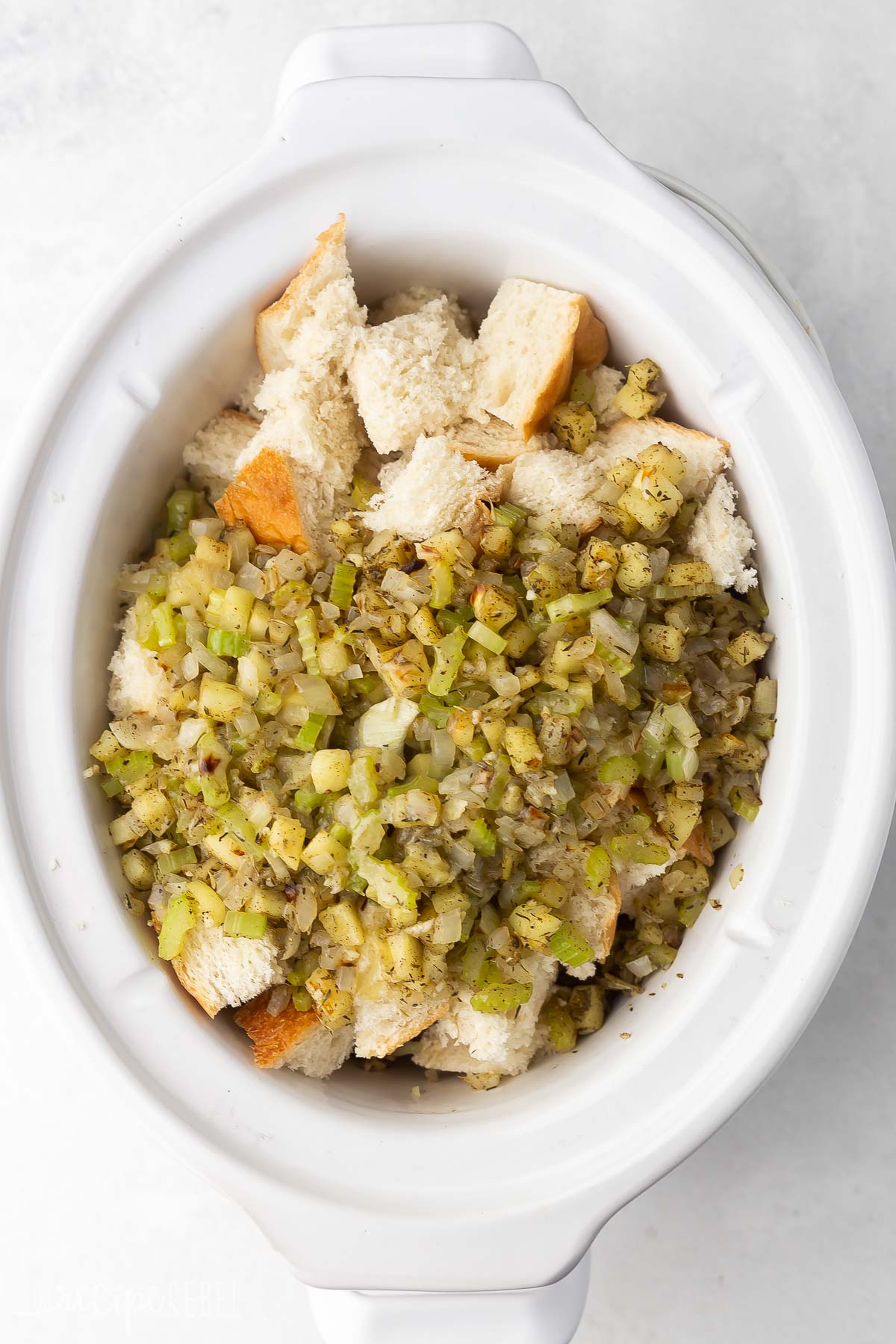 white crockpot with bread cubes in and sauteed vegetables added on top.