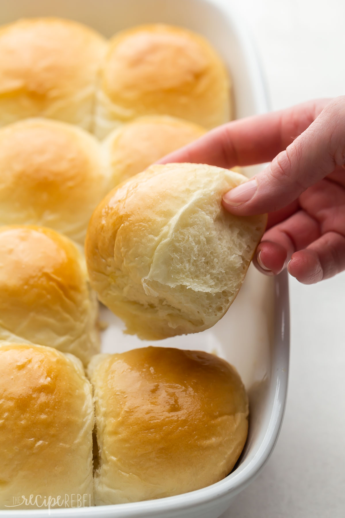 a hand lifting a homemade dinner roll out of white baking dish.